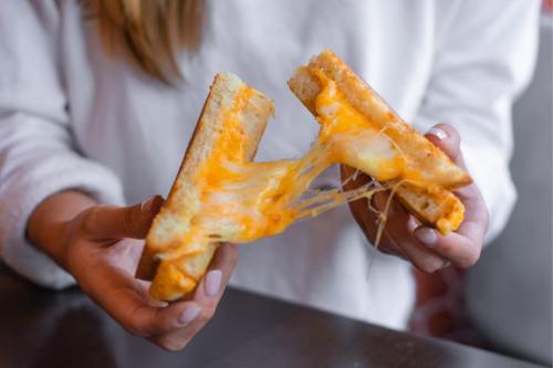 A person holds a gooey grilled cheese sandwich, with melted cheese stretching between two slices of toasted bread.