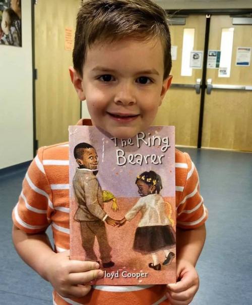 A young boy in an orange striped shirt holds the book "The Ring Bearer" by Lloyd Cooper, smiling at the camera.