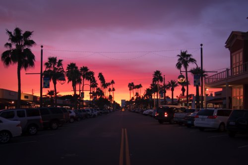 A vibrant sunset casts pink and purple hues over a palm-lined street filled with parked cars.