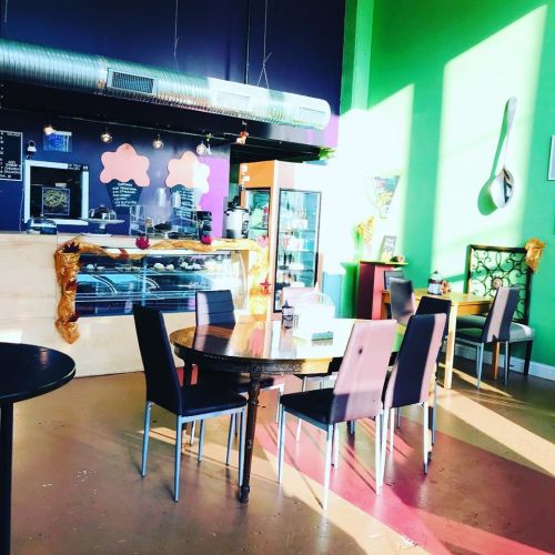 Bright café interior with green and purple walls, a display case, and tables set for customers. Sunlight streams in.
