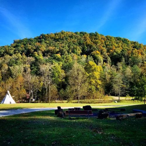 A scenic view of a green hill with autumn foliage, featuring a teepee and benches in a grassy area below.