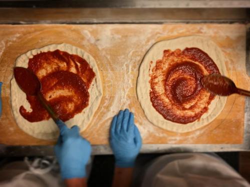 Two hands in blue gloves spread tomato sauce on pizza dough on a floured surface.