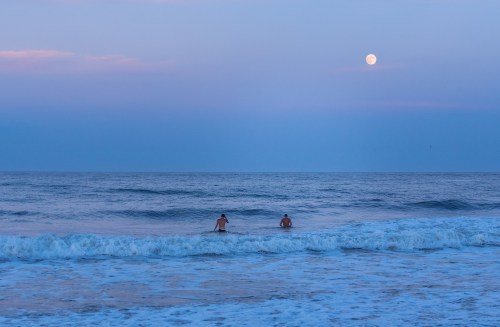 Two people wade into the ocean at dusk, with a full moon glowing in the sky above the calm waters.