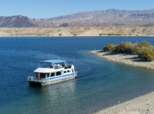 A houseboat navigates a calm lake surrounded by sandy shores and mountains in the background.