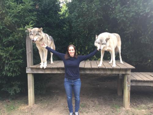 A woman stands on a wooden platform with two wolves, one on each side, surrounded by greenery.