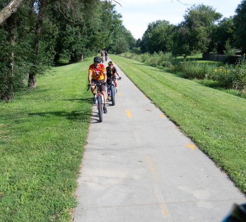 A group of cyclists riding on a paved path surrounded by greenery and trees on a sunny day.