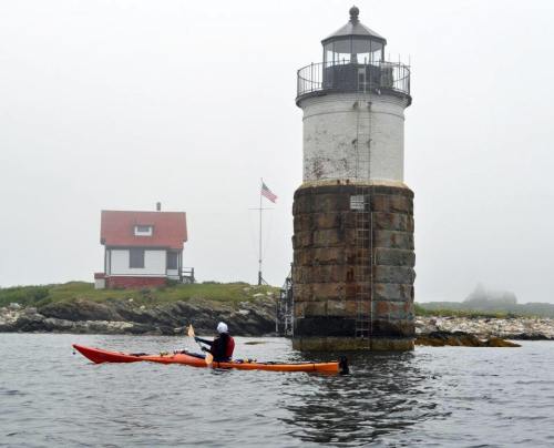 A kayaker paddles near a foggy lighthouse and a red-roofed house on a rocky shore.
