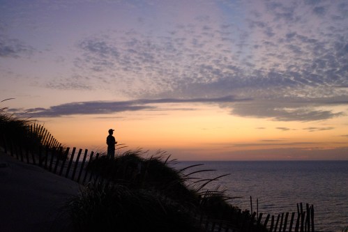 A person stands on a dune at sunset, overlooking the ocean with a colorful sky and silhouetted grass in the foreground.