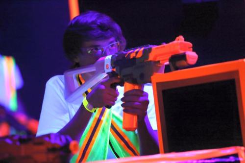 A person aiming a toy gun in a neon-lit environment, wearing a white shirt and glowing accessories.