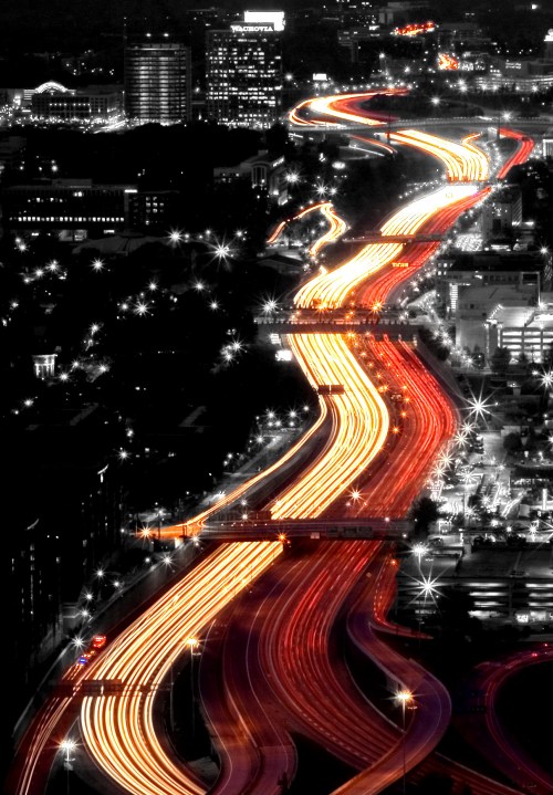 Aerial view of a busy highway at night, with streaks of red and white lights against a black and white cityscape.