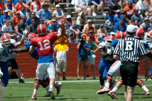 A football player in a red jersey throws a pass while defenders rush towards him; spectators watch in the background.