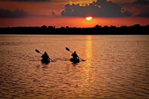 Two kayakers paddle across a shimmering lake at sunset, with vibrant clouds and a glowing sun in the background.