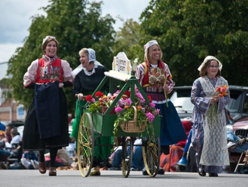 Four women in traditional attire walk together, smiling, with a flower cart in a festive outdoor setting.