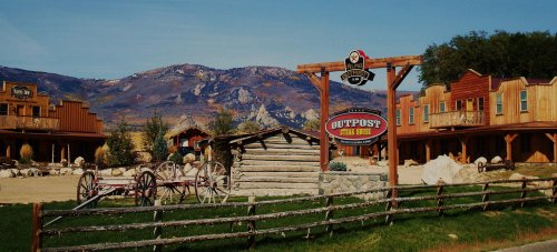 A rustic western outpost with wooden buildings, a sign, and mountains in the background under a clear blue sky.