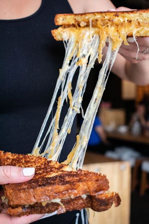 A person holds a grilled cheese sandwich, with melted cheese stretching between two golden-brown slices of bread.