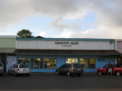 A bakery storefront with a sign reading "Hanemitsu's Bakery & Coffee Shop," featuring parked cars and a cloudy sky.