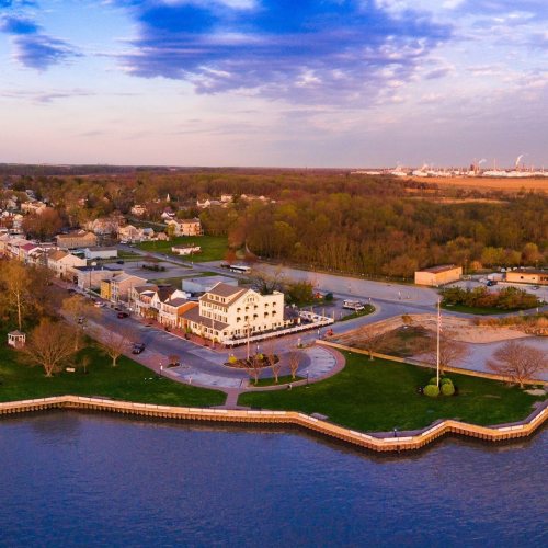 Aerial view of a riverside town with green parks, buildings, and a winding waterfront under a colorful sky.