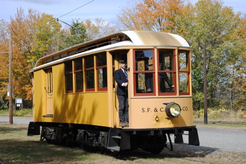 A vintage yellow streetcar with a conductor standing at the front, surrounded by autumn trees.