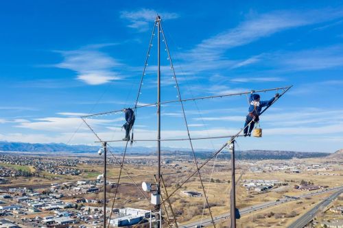 Two workers climb a tall metal star structure against a clear blue sky, overlooking a valley and distant mountains.