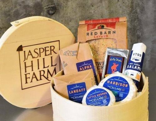 A wooden box labeled "Jasper Hill Farm" filled with various cheeses and snacks, including salami and lavash.