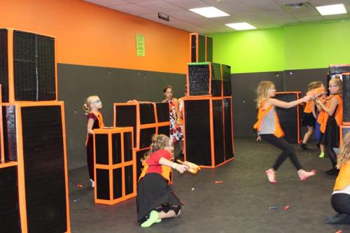 Children playing in a colorful indoor arena, using foam blasters and obstacles for a fun, active game.