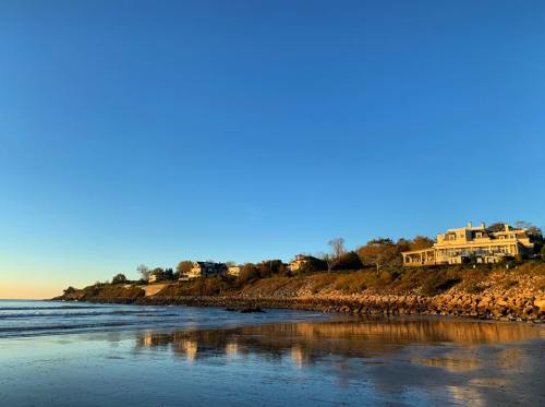 Coastal view with sandy beach, rocky shore, and houses on a hillside under a clear blue sky.
