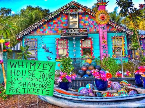 Colorful, whimsical house with vibrant decorations and a sign for a house tour. Flowers and painted objects adorn the yard.