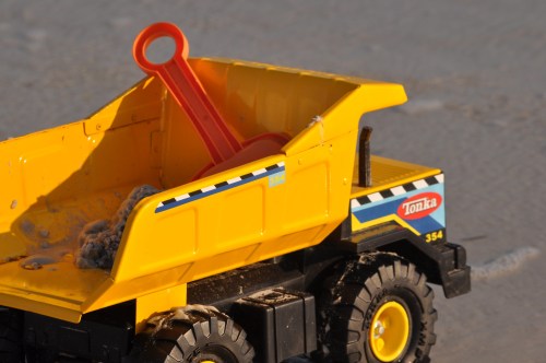 A yellow Tonka toy dump truck with a red shovel in its bed, sitting on a sandy surface.
