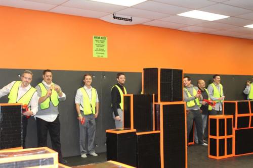 A group of men in safety vests stand behind black and orange foam barriers, holding toy guns in a play area.