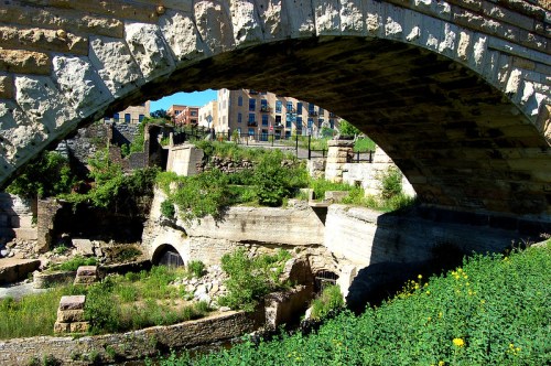 A stone arch bridge spans over a lush, green area with ruins and buildings in the background under clear blue skies.