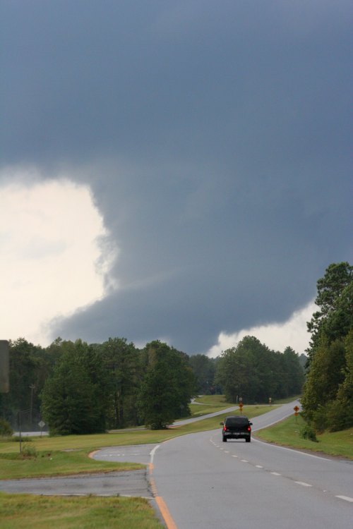 A dark, ominous cloud looms over a winding road, with trees lining the sides and a car driving away.