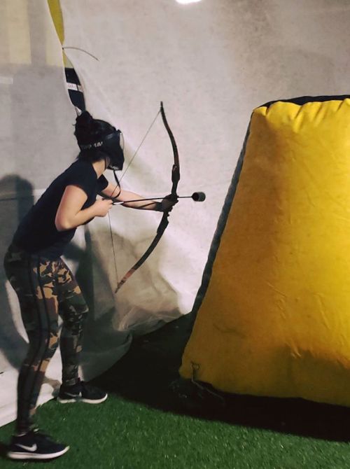 A person wearing a headset aims a bow and arrow at a target behind a large yellow obstacle in an indoor setting.