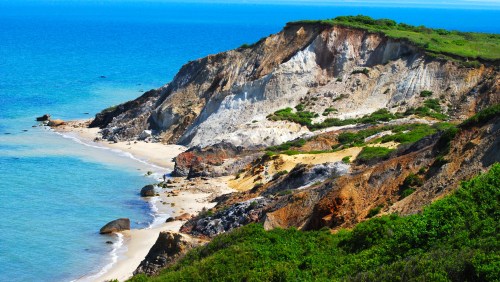 A scenic coastal view featuring cliffs, sandy beach, and clear blue water under a bright sky.