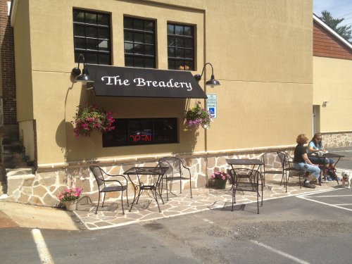 A quaint bakery with outdoor seating, flowers, and an "Open" sign, set in a charming building.