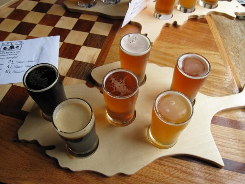A wooden flight board holds six small glasses of different beers, arranged on a table with a chessboard in the background.