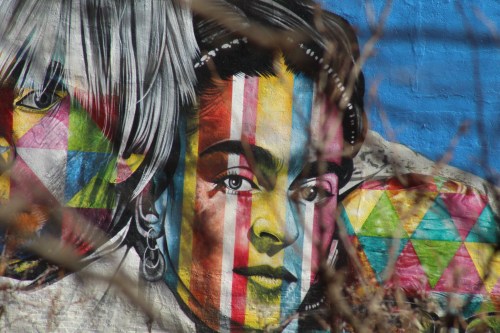 Colorful mural featuring a woman's face with geometric patterns and vibrant stripes, partially obscured by branches.