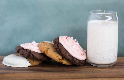 Two large cookies with pink frosting and crushed peppermint, next to a glass of milk on a wooden surface.