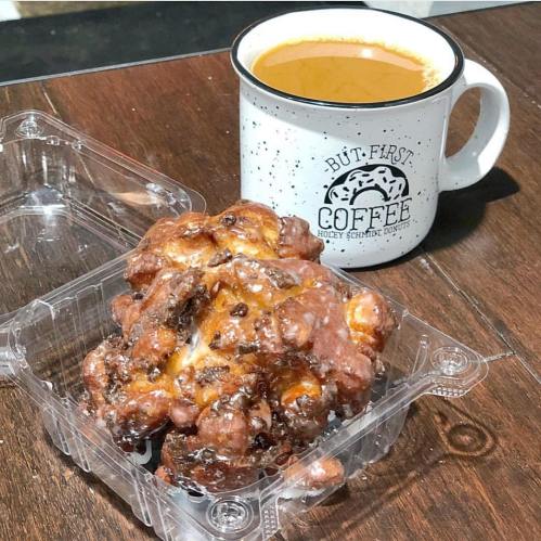 A coffee mug beside a container holding a glazed apple fritter on a wooden table.