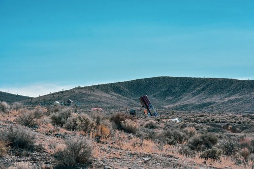 A hilly desert landscape with sparse vegetation and a tilted structure in the foreground against a clear blue sky.