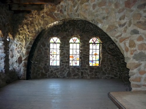 Interior of a stone room featuring three arched stained glass windows and a wooden floor.
