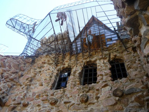 A stone building with a metal cage structure above, featuring figures and a wooden roof, under a clear blue sky.