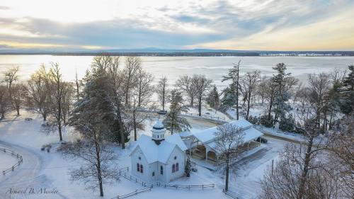 A snowy landscape featuring a small white church and trees, with a frozen lake and cloudy sky in the background.