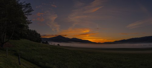 A serene landscape at sunset, featuring mountains, a misty valley, and colorful clouds in the sky.