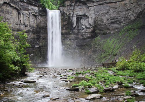 A tall waterfall cascades down rocky cliffs, surrounded by lush greenery and a river with people observing nearby.