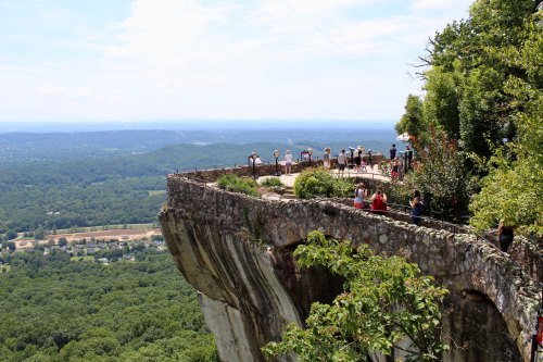 A scenic overlook with people taking photos on a rocky ledge, surrounded by lush green hills and a clear blue sky.