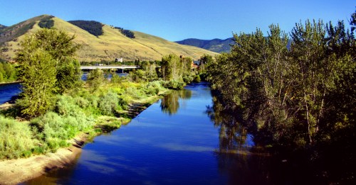 A serene river flows through lush greenery, with rolling hills and a clear blue sky in the background.
