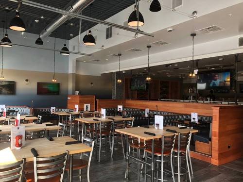 Modern restaurant interior with wooden tables, metal chairs, and a bar area, featuring menus and a TV in the background.