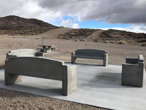 Concrete benches arranged in a circle on a paved area, with a desert landscape and cloudy sky in the background.