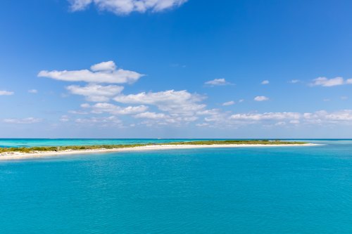 A serene coastal view featuring turquoise waters, a sandy beach, and a clear blue sky with scattered clouds.