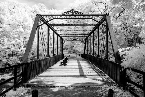 A black and white image of an old iron bridge surrounded by trees, casting shadows on the wooden walkway.